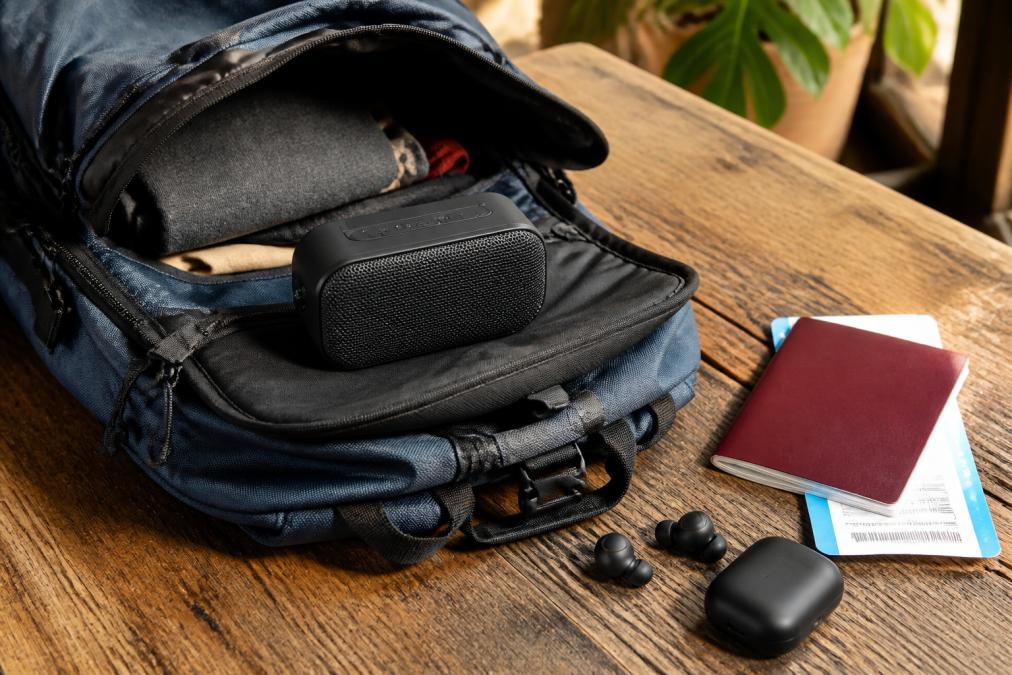 A compact Bluetooth speaker resting on top of an open travel backpack on a rustic wooden table, with a passport, boarding pass, and earbuds arranged nearby in warm natural daylight