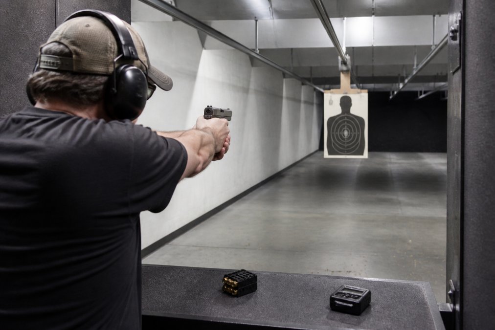 A beginner shooter at an indoor range practicing a controlled pair drill on a paper target, with a shot timer on the bench and a speed loader nearby