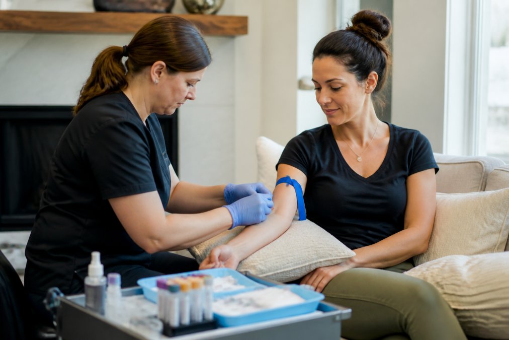 A professional phlebotomist in a clean home setting gently drawing blood from a relaxed patient seated on a couch, with sterile equipment arranged on a nearby tray, soft natural window light