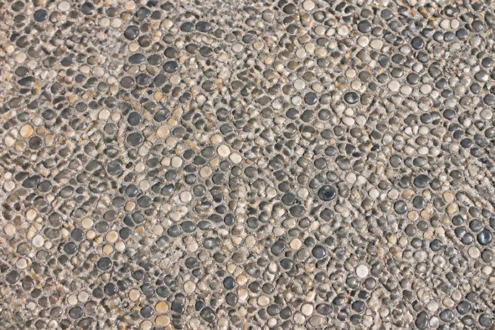 Close-up detail of exposed aggregate concrete surface showing visible rounded pebbles and stones in shades of gray and tan with natural texture and color variation under bright sunlight