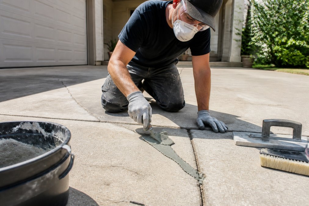 Professional tradesperson using a trowel to apply concrete resurfacing compound into cracks on a driveway on a sunny day, with proper safety equipment visible