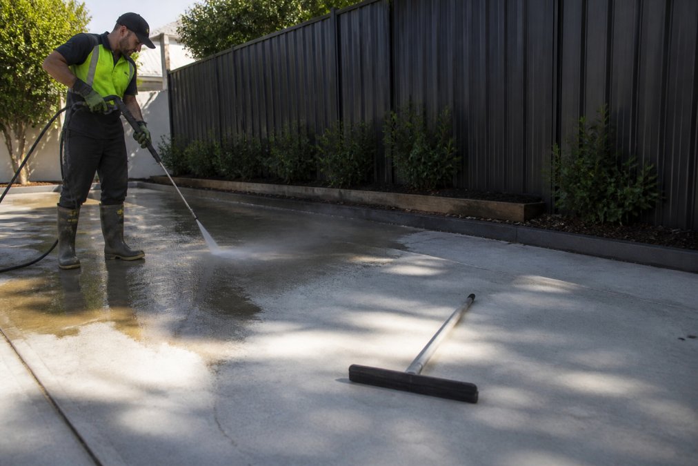 A worker using a high-pressure washer to clean a concrete driveway, water spraying off the surface with a squeegee resting nearby in bright daylight