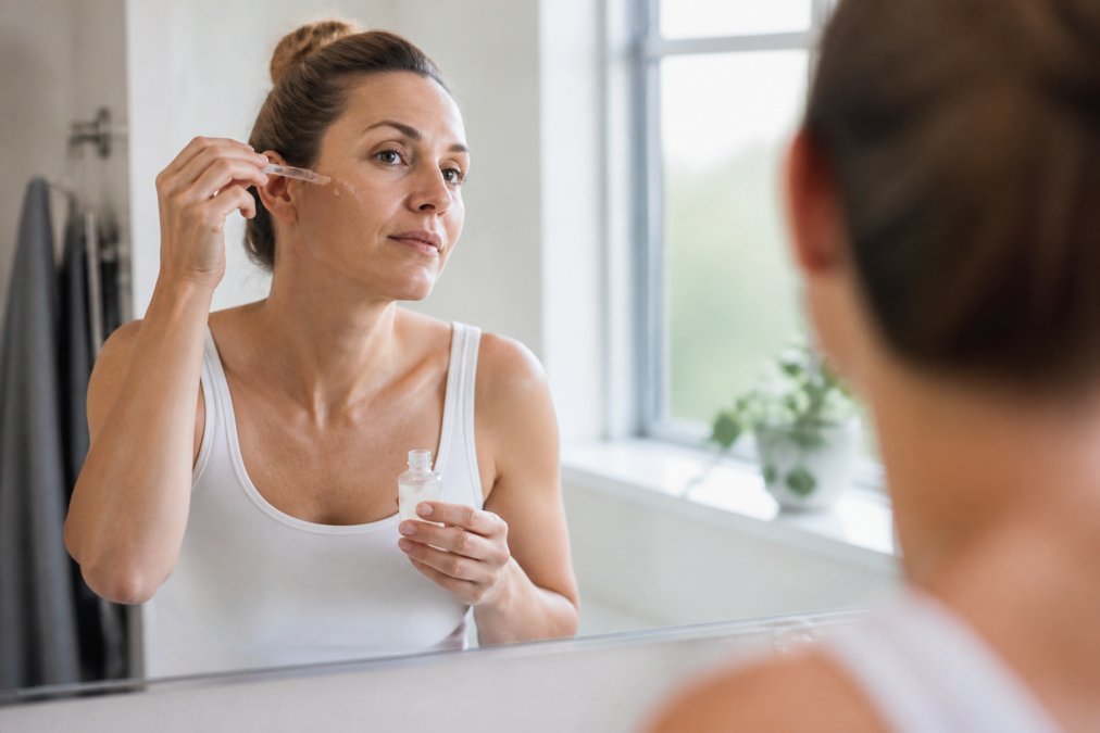 Woman applying copper peptide serum from a dropper in a bright bathroom mirror