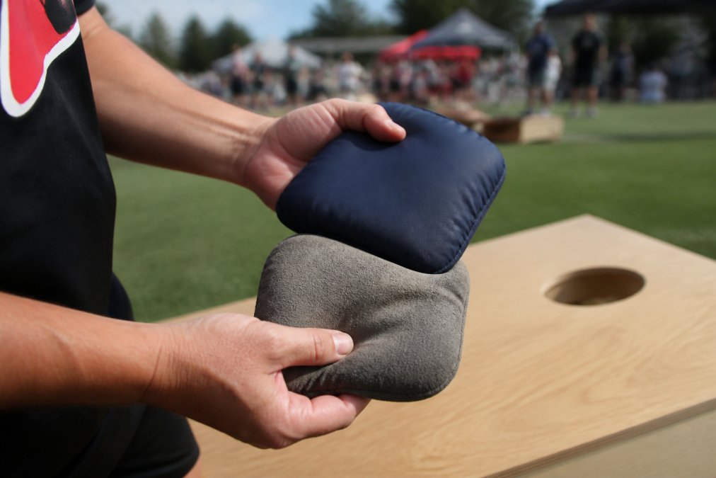Close-up of a player's hands holding two dual-sided cornhole bags over a wooden board, one side showing suede texture and the other smooth synthetic fabric, with a blurred tournament setup in the background