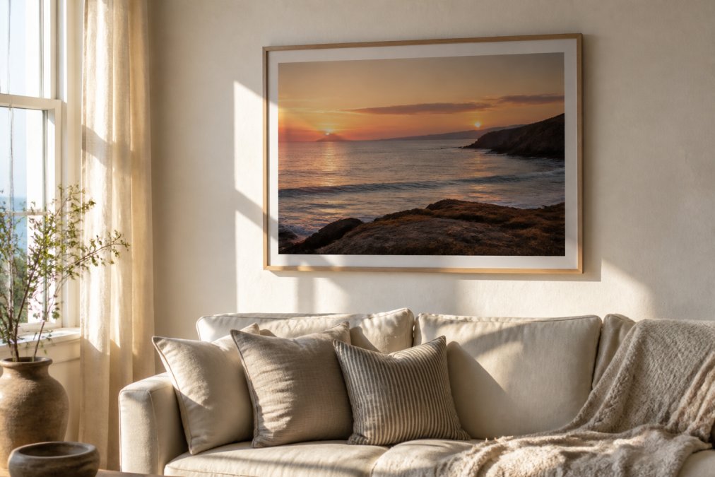 A sunlit living room wall displaying a framed coastal art print of a Cornish sunset, with a linen sofa and soft neutral textiles below