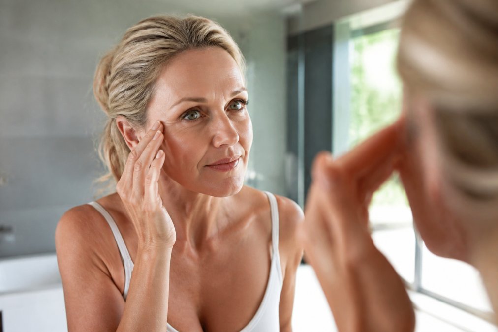 Close-up of a woman in her 50s looking in a mirror, gently touching the fine lines around her eyes, in a bright, modern bathroom