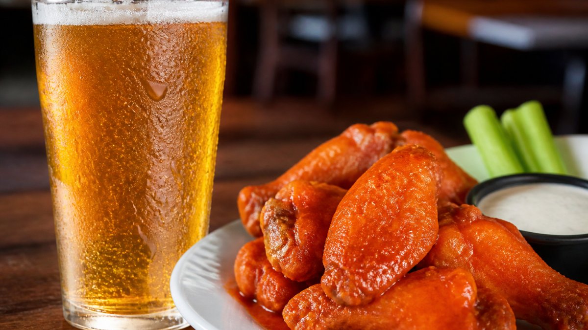 Cold IPA glass with condensation beading on the surface, positioned next to a plate of traditional buffalo wings with tangy sauce glistening under warm lighting, close-up detail showing both items