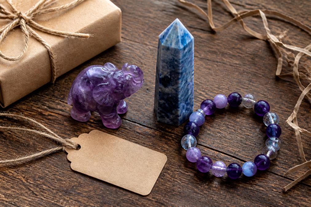 Close-up overhead shot of a curated crystal gift set arranged on a rustic wooden table, featuring a purple amethyst elephant carving, a polished blue tower, and a beaded bracelet beside a handwritten gift tag and kraft paper wrapping in soft natural window light