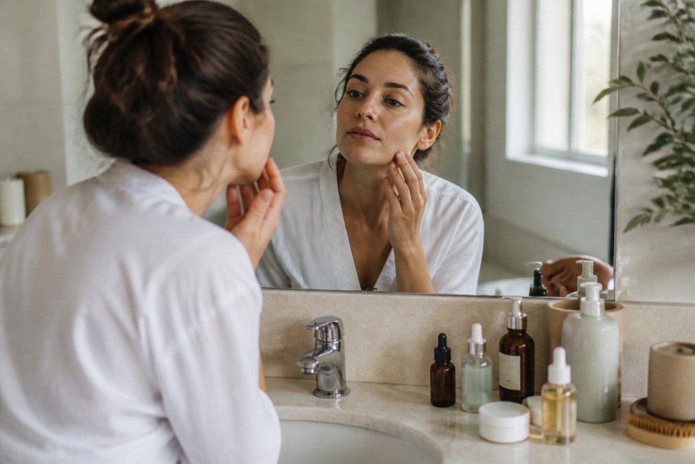 Woman examining her skin in a mirror surrounded by curated beauty essentials on a bright bathroom counter.