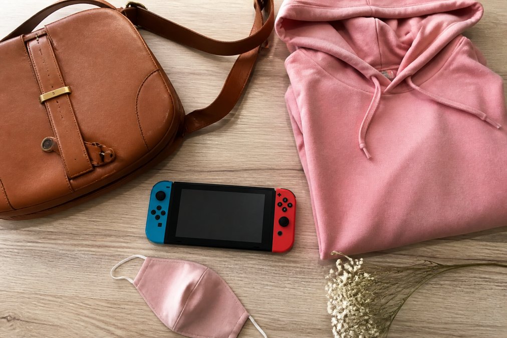 A flat-lay photograph of curated lifestyle products arranged on a light wooden surface, including a leather handbag, a pink hoodie, a portable game console, and a face mask, styled like a social media product haul
