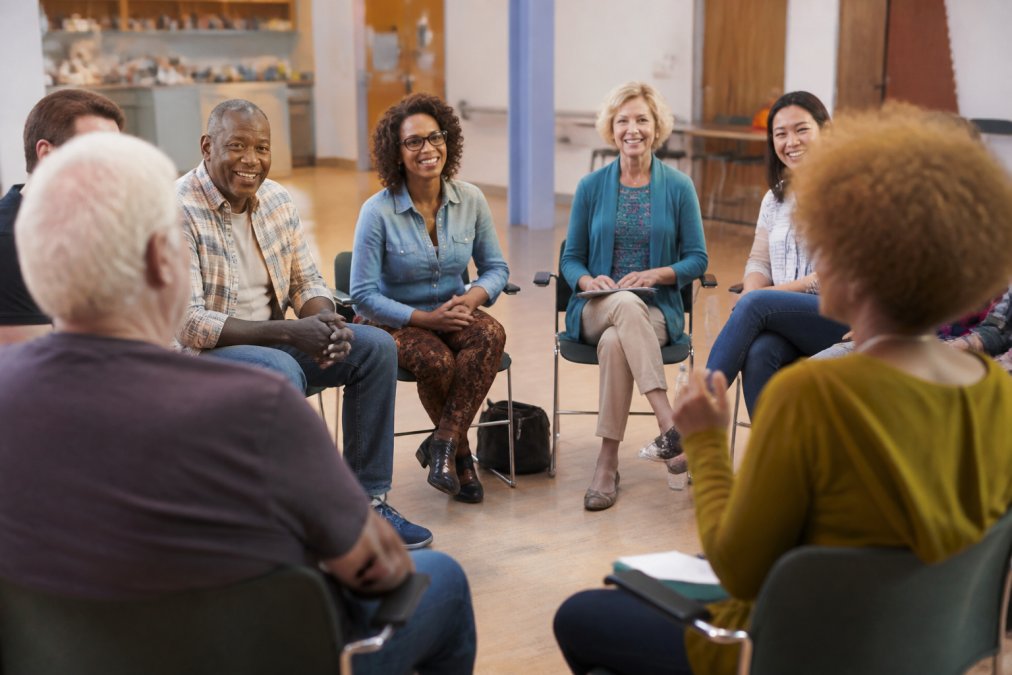 A diverse group of people sitting in a circle during a workshop or community gathering, engaged in discussion, showing genuine connection and support