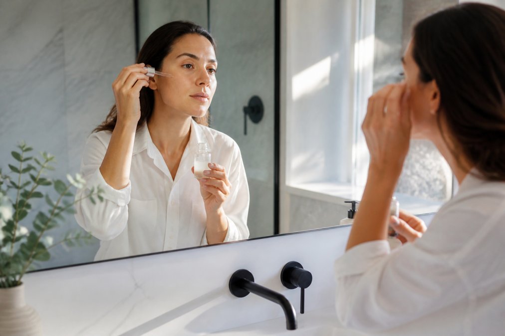 Professional woman in minimalist bathroom applying lightweight serum to face at mirror during morning routine, natural window lighting streaming across modern vanity