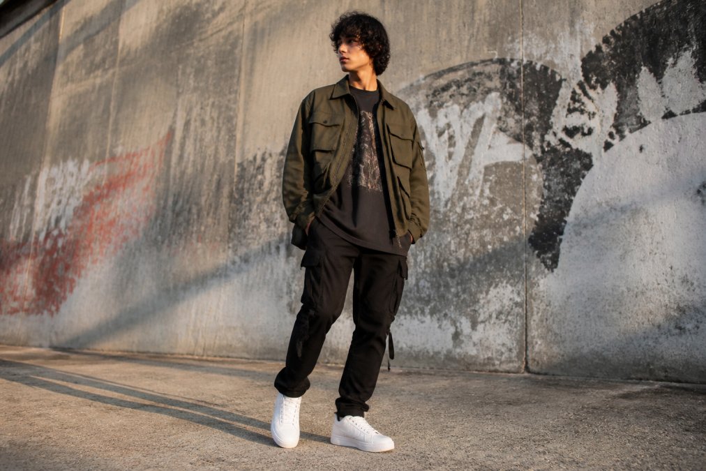 A fashion-forward young adult wearing an oversized graphic tee layered with a utility jacket and clean white sneakers, posed against an urban concrete wall with faded graffiti, shot in natural afternoon light