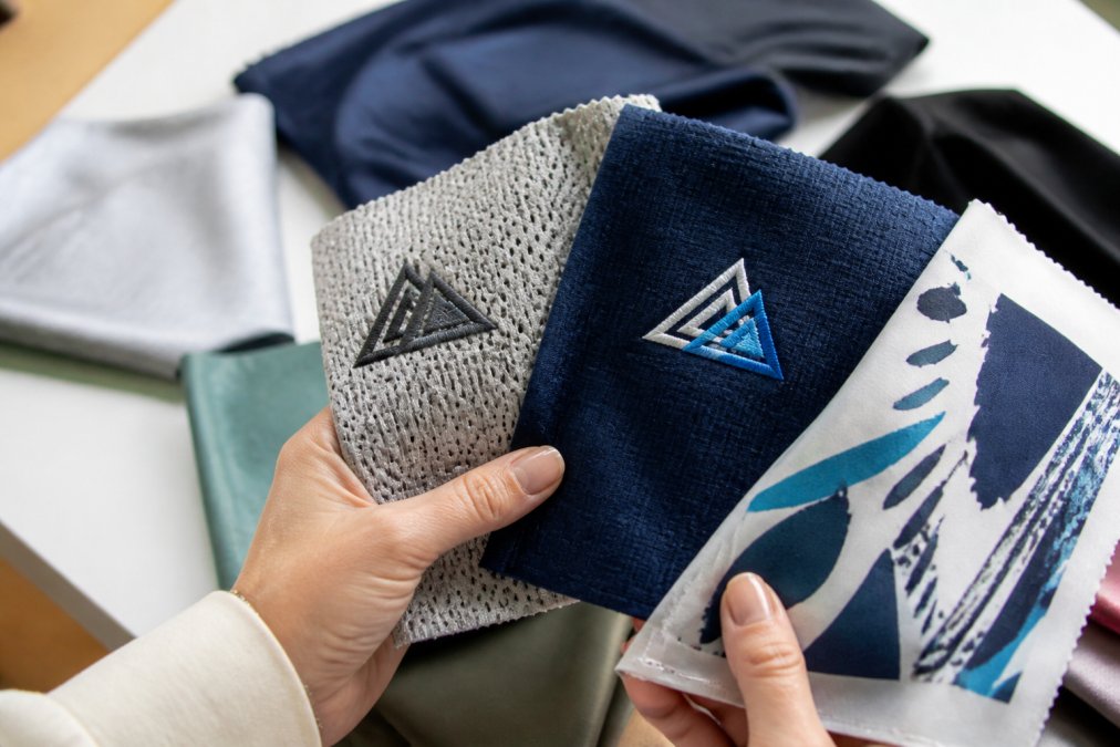 Close-up of hands holding custom apparel swatches showing different performance fabric textures, with visible embroidery and direct-to-film print details in natural studio lighting