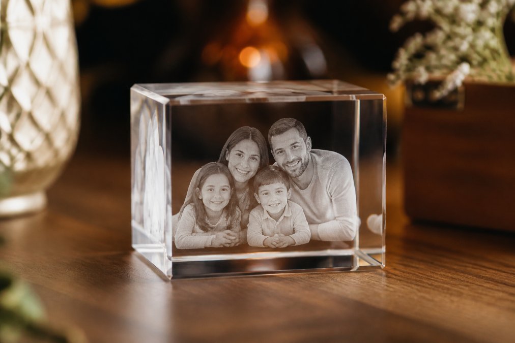 Close-up of a 3D crystal cube displaying a family photo engraved inside with remarkable depth and clarity, sitting on a polished wooden surface with soft warm lighting highlighting the crystal's refractive qualities
