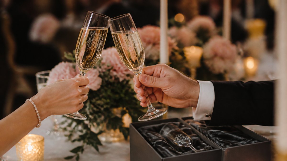 Close-up of two engraved champagne flutes being raised in a toast at an elegant wedding reception, soft candlelight and floral centrepieces blurred in the background