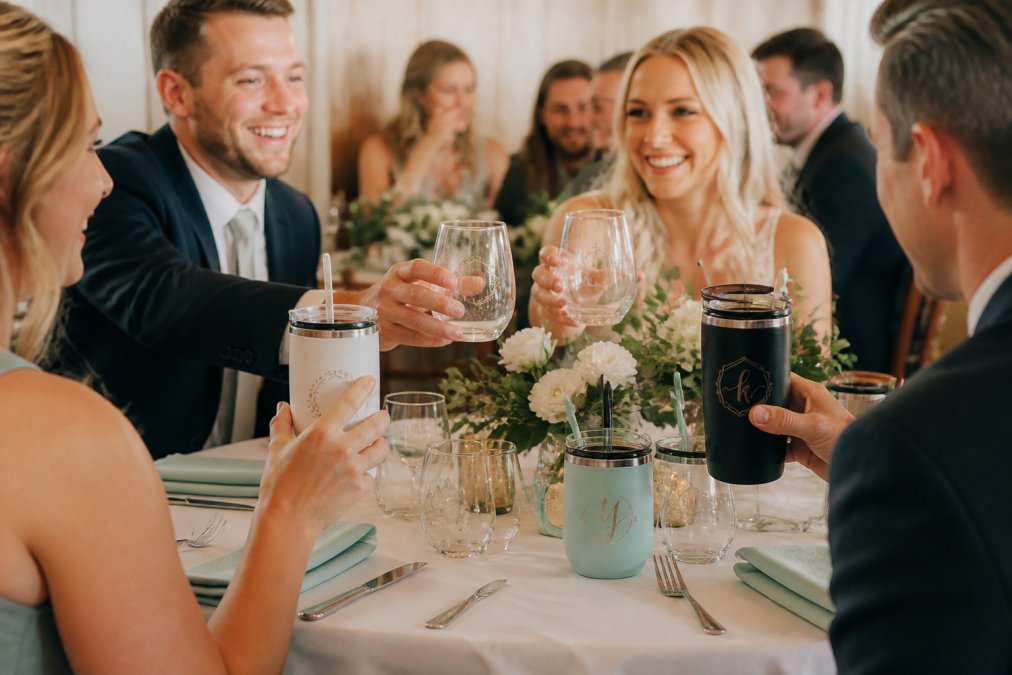 A table at a wedding reception set with an assortment of personalized drinkware favors, including stemless wine glasses and acrylic tumblers with engraved names and dates, soft natural light, floral centerpiece in the background