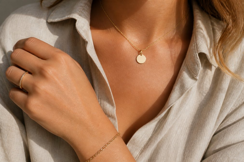 A flat-lay photo of a woman's wrist and neck wearing a dainty gold necklace with a small engraved disc pendant and a thin stackable ring, styled with a neutral linen shirt and soft natural light
