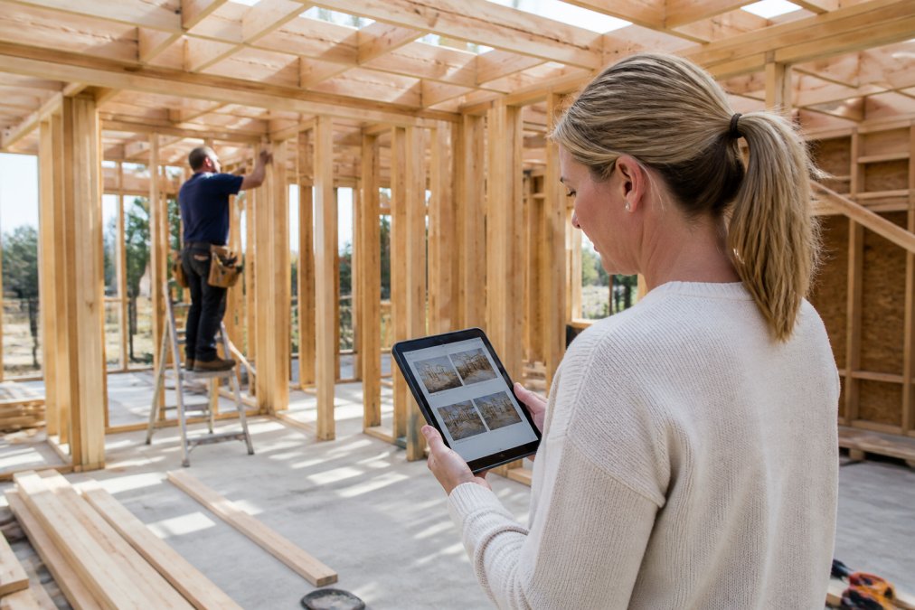 Homeowner reviewing project updates on a tablet while standing in a construction site, with contractor visible in the background working on framing