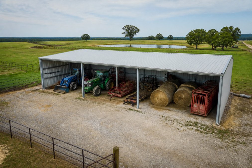 Aerial view of a rural farm property showing a modern galvanized steel storage shelter protecting tractors, hay bales, and livestock equipment from rain and sun exposure, with fencing and pastures visible in the background