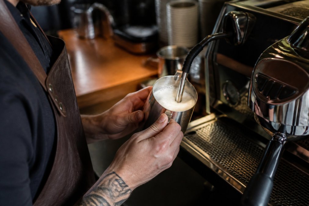 Close-up of a barista's hands holding a stainless steel pitcher, steam wand submerged in milk with visible microfoam texture, stainless steel espresso machine in the background, warm cafe lighting
