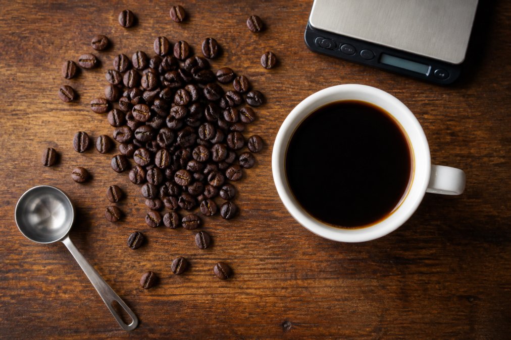 Overhead shot of dark roasted coffee beans scattered next to a freshly brewed cup of black coffee on a wooden table, with a small digital scale and measuring spoon nearby