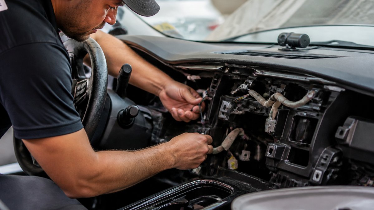 Professional technician with precision tools installing hardwired dash camera behind vehicle dashboard, carefully routing cables through existing wire channels and connecting to vehicle's electrical system