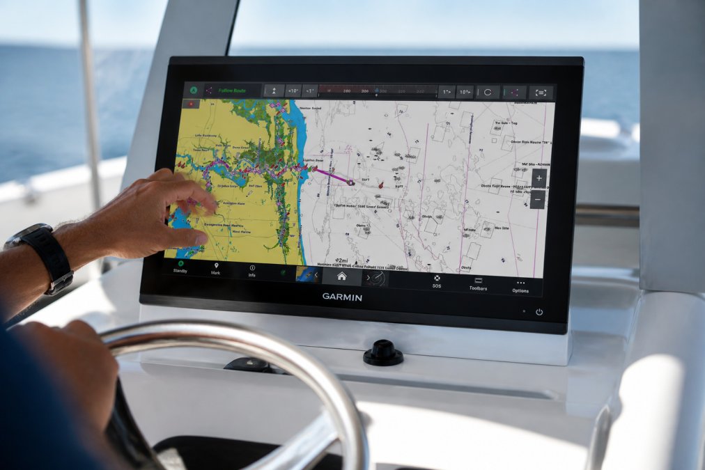 Professional boat captain's hands adjusting controls on a fully integrated Garmin chartplotter display mounted on a modern helm, showing real-time navigation data, sonar readouts, and touchscreen interface with natural sunlight reflecting off the screen