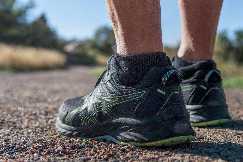 Close-up of a trail runner's foot on rocky terrain, showing a high-cut debris-shielding sock with reinforced heel and protective mesh cuff visible against rough ground and scattered stones