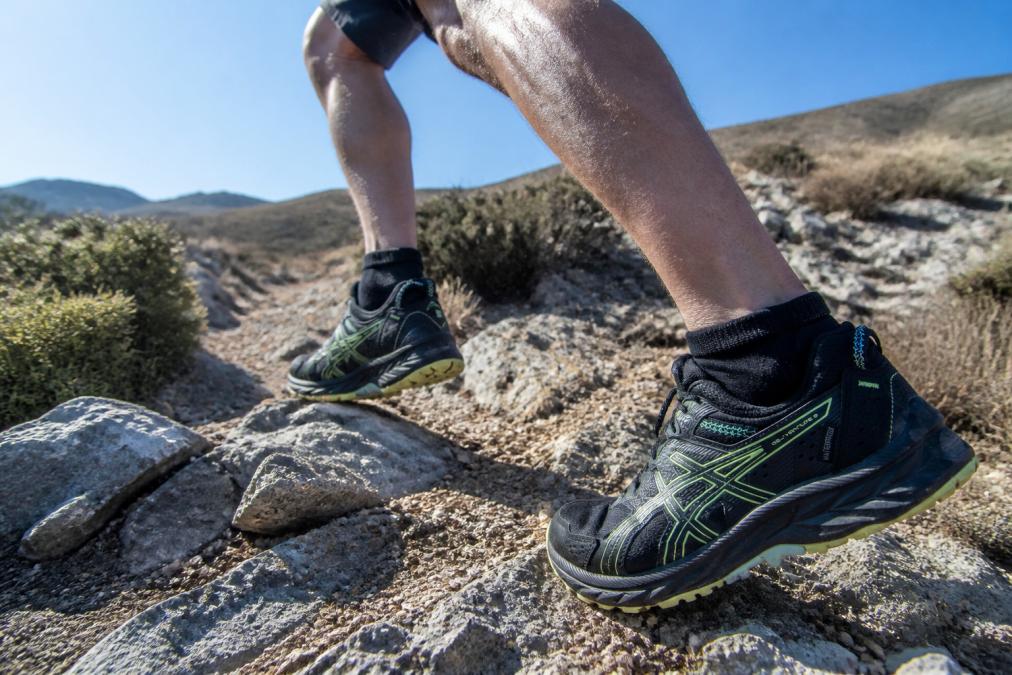 Outdoor action shot of a trail runner in motion on technical rocky terrain, wearing high-cut socks with visible integrated gaiter cuff, demonstrating how the protective design shields against sand, grit, and debris exposure
