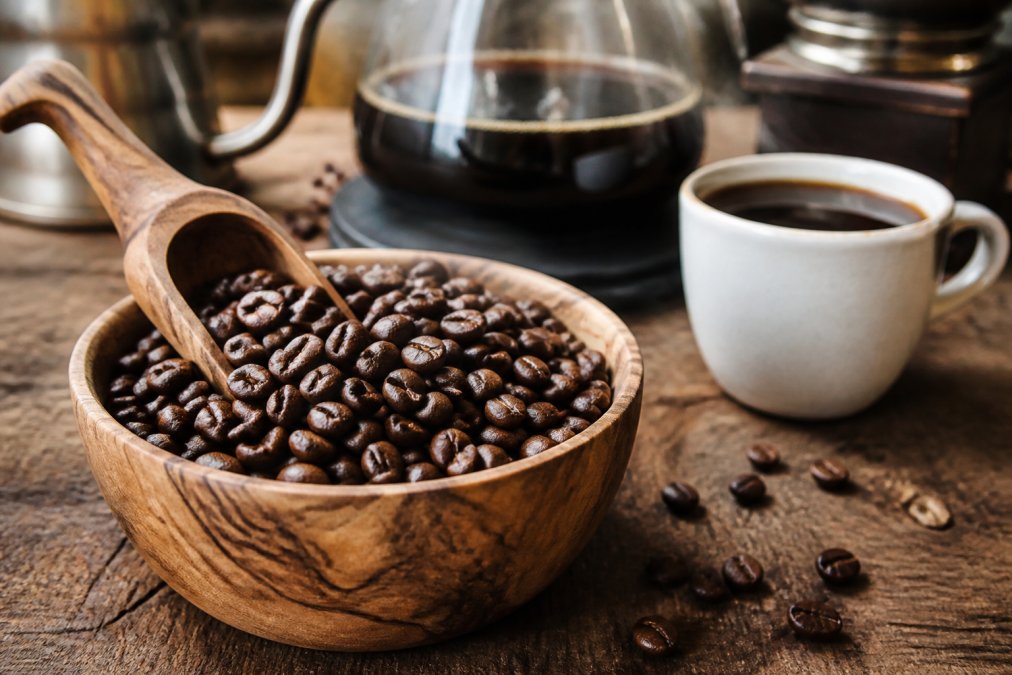 A close-up of freshly roasted decaf coffee beans in a wooden bowl with a scoop, a cup of black coffee and brewing equipment softly blurred in the background