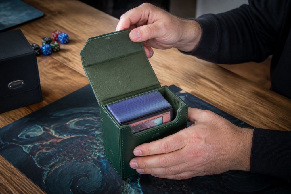 A tabletop gamer's hands opening a magnetic-closure deck box filled with double-sleeved trading cards, with a playmat, dice, and a Commander deck box nearby on a wooden table