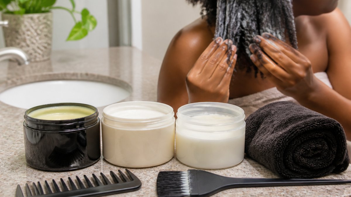 A flat lay of three deep conditioner jars with a hair mask brush, a wide-tooth comb, and a microfiber towel on a bathroom counter, with a woman's hands applying conditioner to her coily hair in the background