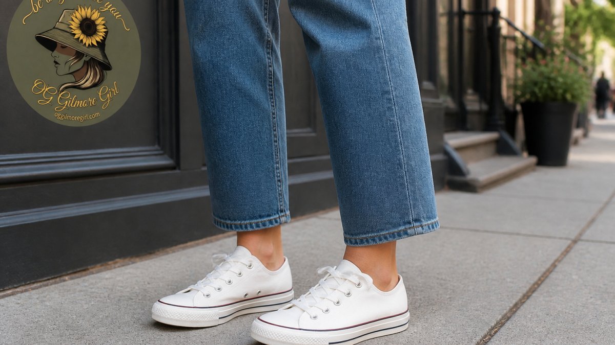 A woman in her late twenties standing on a city sidewalk, viewed from the knees down, wearing straight-leg blue jeans and white canvas sneakers, with the hem sitting just above the shoe so the ankle and sneaker collar are visible