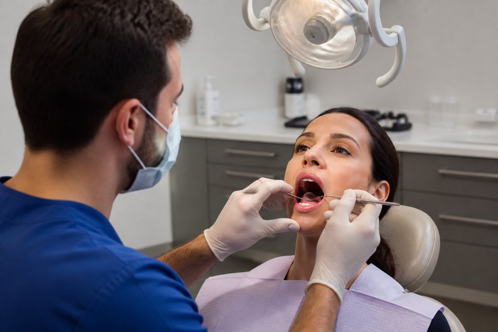 A dentist in blue scrubs examining a patient's open mouth with a dental mirror and overhead light, focusing closely on the soft palate and tongue position during a clinical consultation in a clean, modern dental operatory