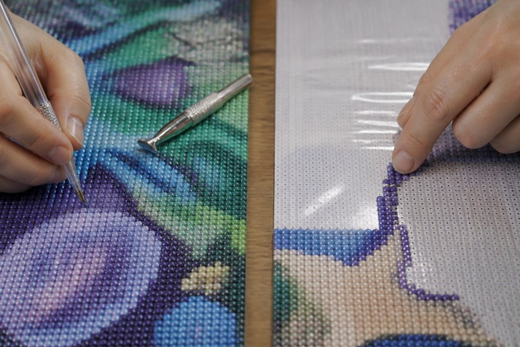 Close-up of a crafter's hands pressing a diamond drill into a glossy poured-glue canvas, with a metal applicator pen resting nearby