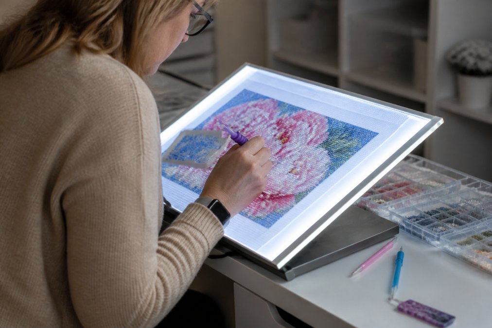 Close-up of hands placing diamond drills on a canvas illuminated by a bright LED light pad, showing proper posture with the pad positioned at a comfortable angle, natural studio lighting