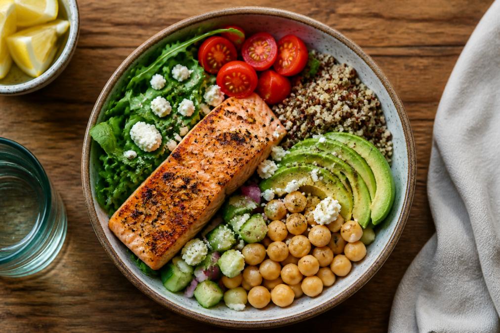 A vibrant, top-down shot of a colorful Mediterranean-style meal bowl with grilled salmon, leafy greens, quinoa, avocado, and chickpeas on a wooden table, natural daylight
