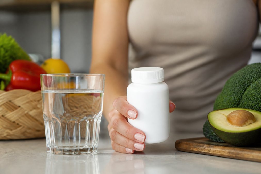 Person holding a bottle of digestive enzymes next to a glass of water on a kitchen counter with fresh produce