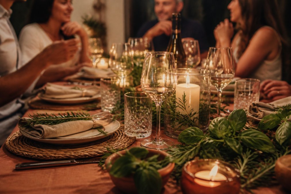 Intimate dinner table set with Italian-inspired decor including candlelit centrepiece, folded linen napkins, wine glasses catching warm light, fresh basil and rosemary sprigs scattered across a terracotta-toned tablecloth, with guests blurred in background enjoying conversation