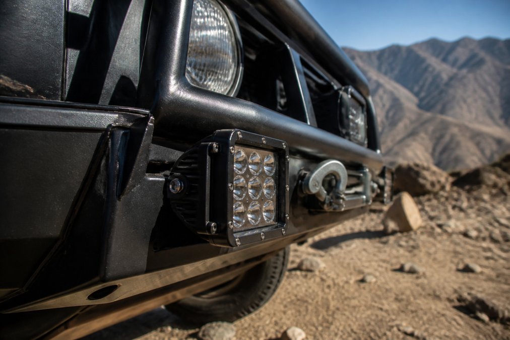 Close-up of a rugged LED off-road light mounted on truck bumper with visible heat sink design and waterproof housing in outdoor terrain