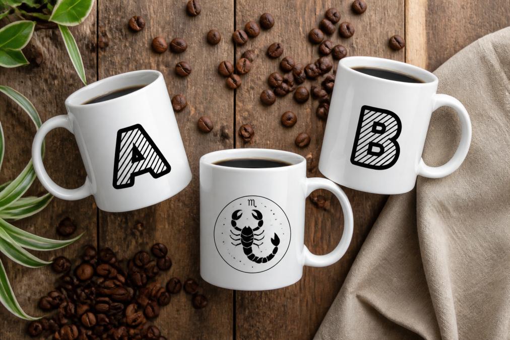 Overhead flat-lay of three personalized ceramic mugs with different alphabet letters and zodiac designs, filled with coffee, arranged on a wooden surface with coffee beans scattered nearby