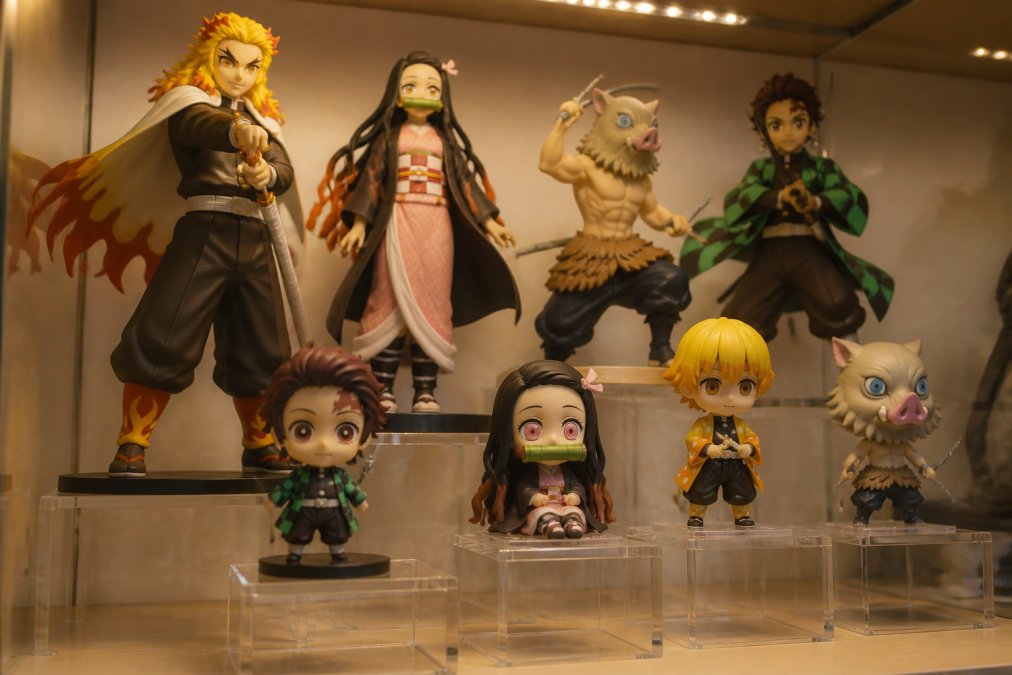 Close-up view of clear acrylic risers at different heights elevating anime figures, including Nendoroids and scale figures, creating visual depth and preventing smaller figures from being hidden behind larger ones, with warm LED lighting from above casting subtle shadows