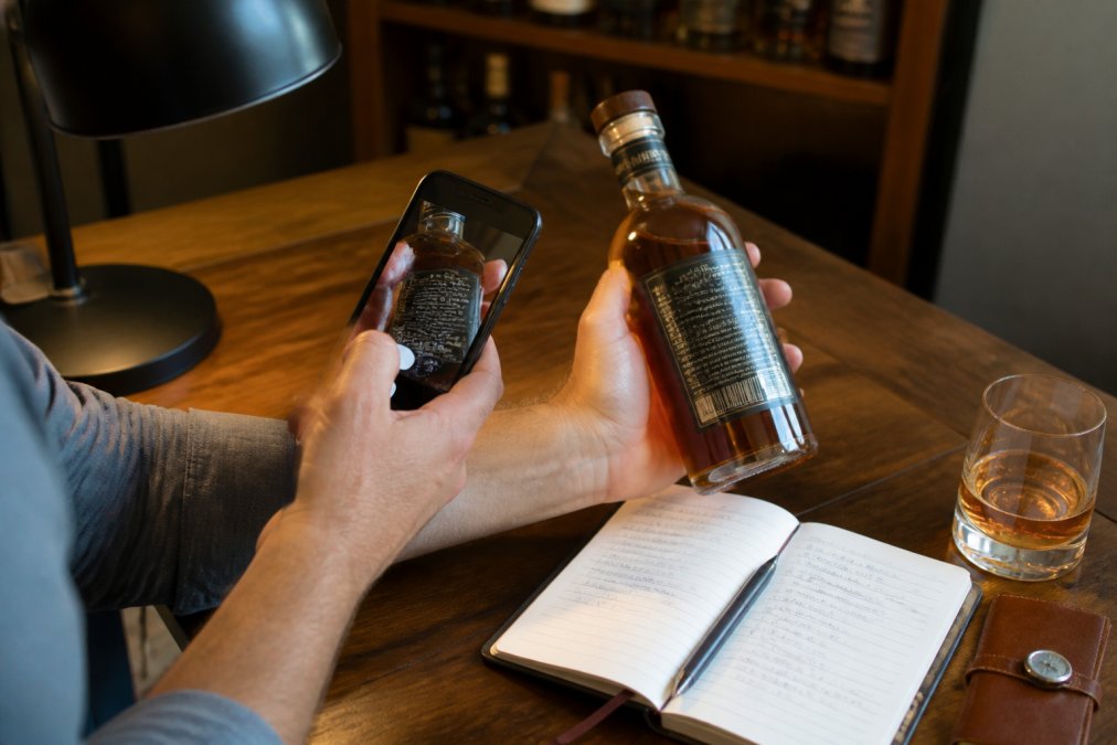 Close-up of hands using mobile app to scan bourbon bottle label with notebook for tasting notes visible nearby, desk lighting