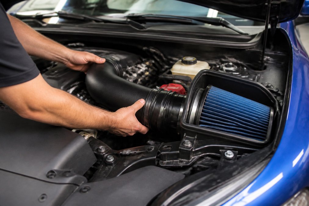 Mechanic's hands installing a cold air intake kit under an open car hood, showing the filter housing and intake tubing connections with engine bay visible