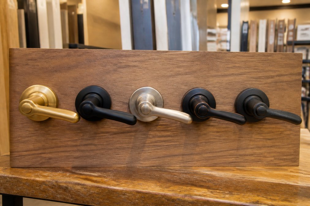 Close-up photograph of five door handles in brass, matte black, satin nickel, oil-rubbed bronze, and powder-coated iron mounted on a wooden display board in a brightly lit showroom