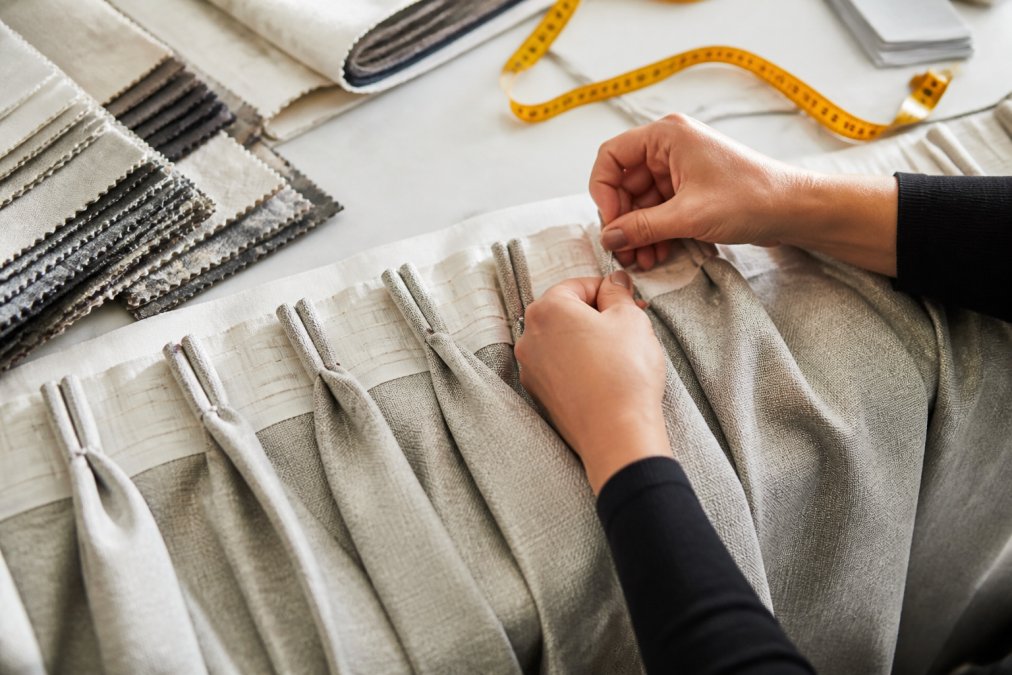 Close-up of hands pinning a pinch pleat header on a drapery panel in a bright workroom, with fabric swatches and measuring tape visible on the table