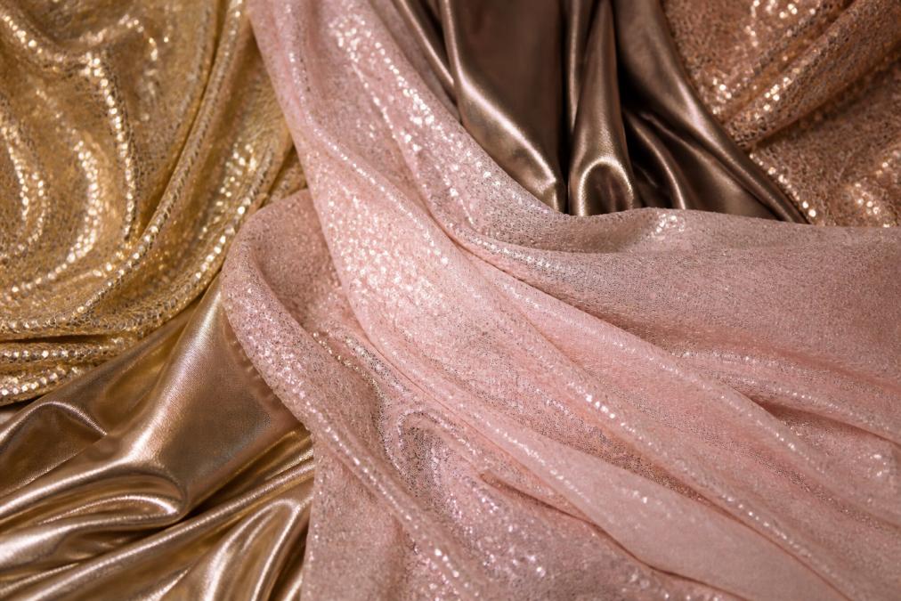 Close-up of luxurious fabrics including sequins, metallic finishes, and satin textures draped elegantly, showing light reflection and shimmer against soft formal lighting