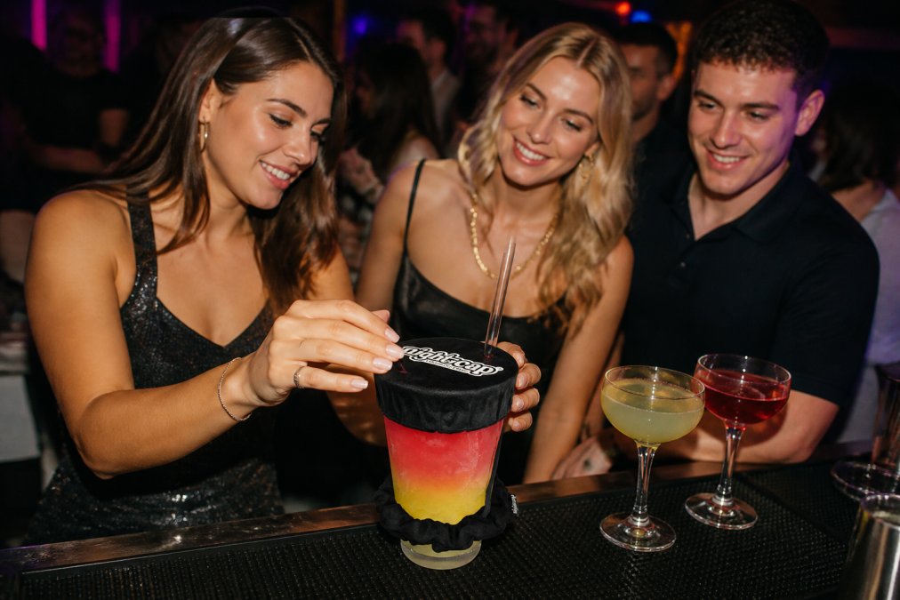 Young adults at a busy club bar, one person placing a drink cover over a glass while friends watch, with vibrant club lighting and neon signs in the background