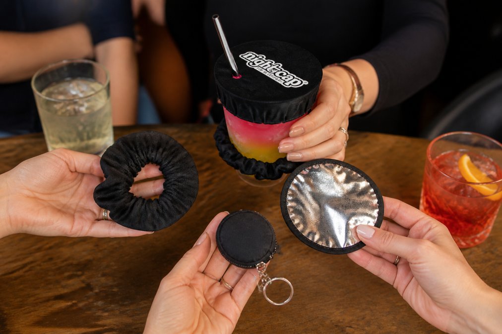 Close-up of hands holding different drink covers displayed on a table: a black scrunchie, a foil lid, and a keychain cover, with drinks nearby in a social setting